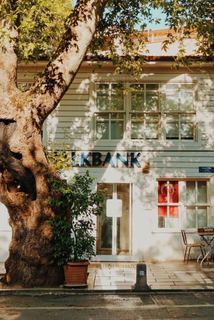 Exterior of a Turkish bank building with white wooden siding, large windows and a tree casting shade.