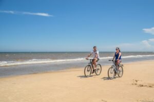 Two people cycling along a sandy beach shoreline, showing eco‑friendly transport options for getting around Kalkan.