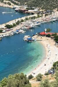Turquoise harbour beach near Kalkan with marina boats, white sun loungers and coastal hillside views.
