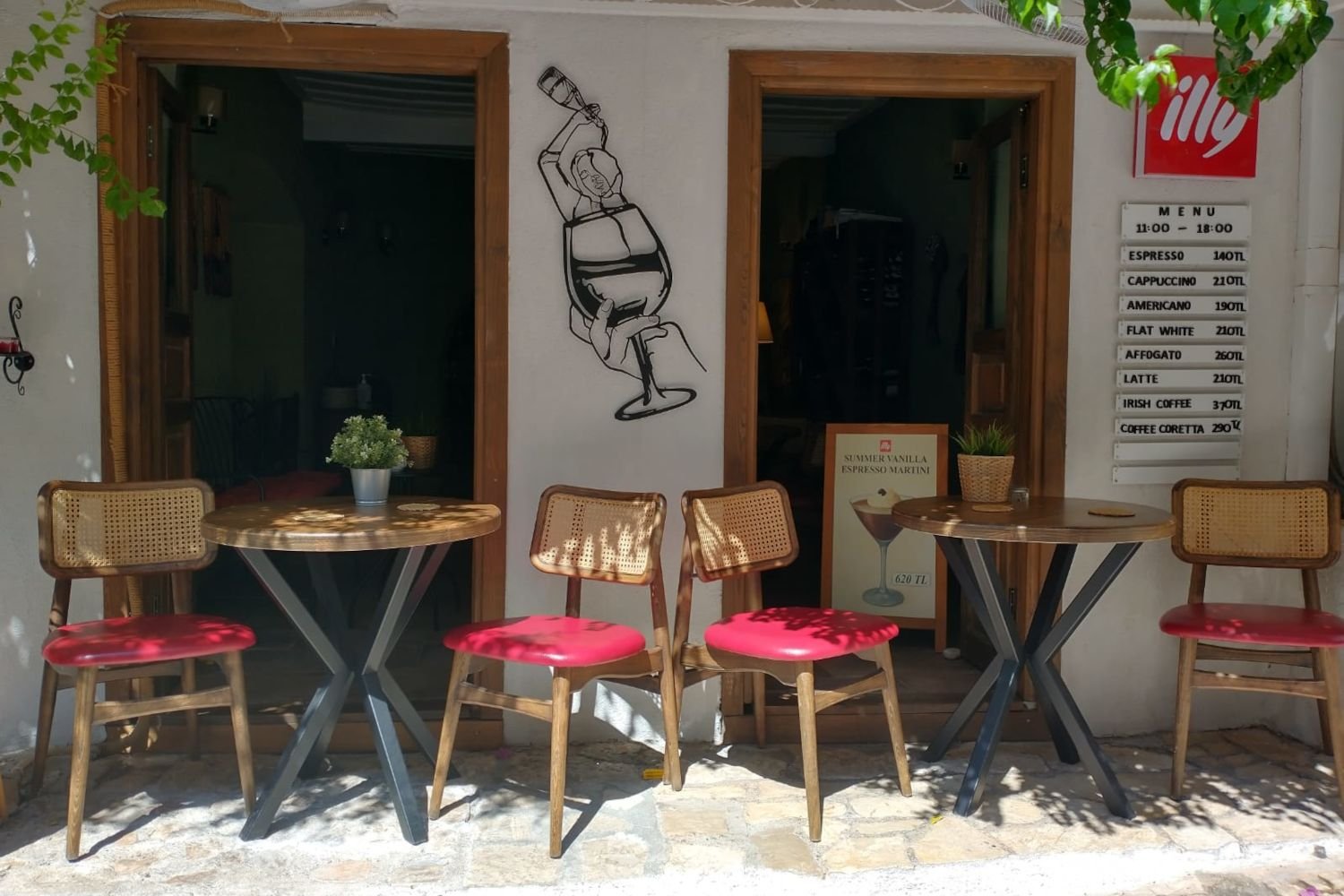 Outdoor seating area of a small café in Kalkan with wooden tables, red‑cushioned chairs, Illy coffee signage and a visible drinks menu.
