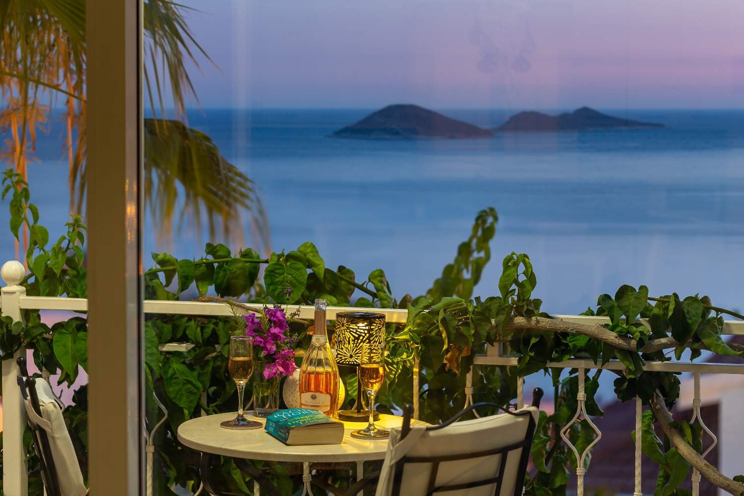 Balcony at Kalkan Limon Apartment with wine glasses, warm lighting and a sea view at sunset.