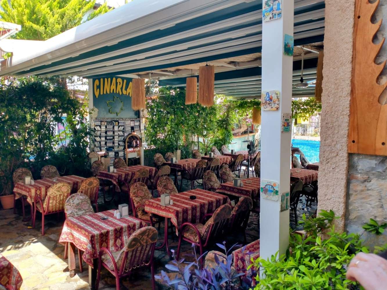 Shaded garden‑style restaurant in Kaş with red patterned tablecloths, cushioned chairs and lush greenery.