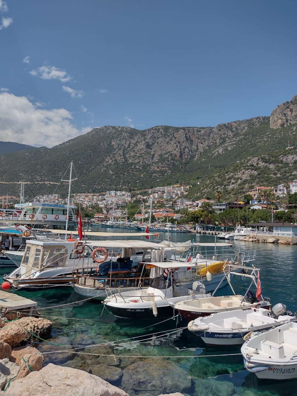 Kaş harbour with small boats, clear turquoise water and hillside town beneath rugged green mountains.