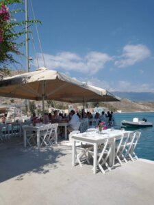 Seaside restaurant in Meis with white tables, umbrellas and colourful waterfront buildings across the bay.