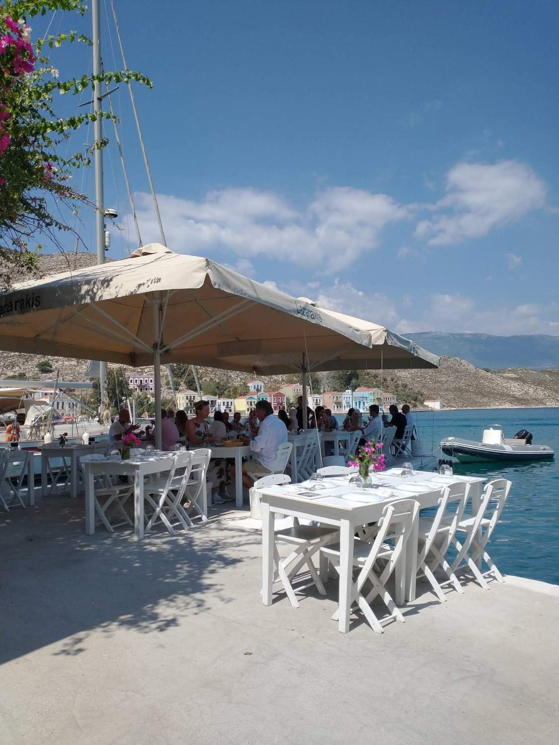 Seaside restaurant in Meis with white tables, umbrellas and colourful waterfront buildings across the bay.